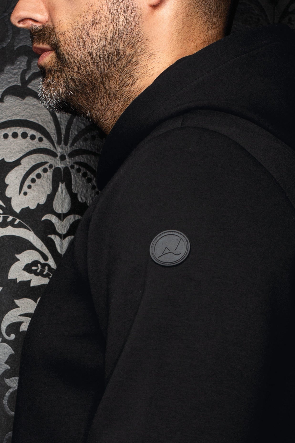 Men's side close-up of black hooded zip sport jacket showing raglan seam and tonal circular sleeve logo patch by Au Noir